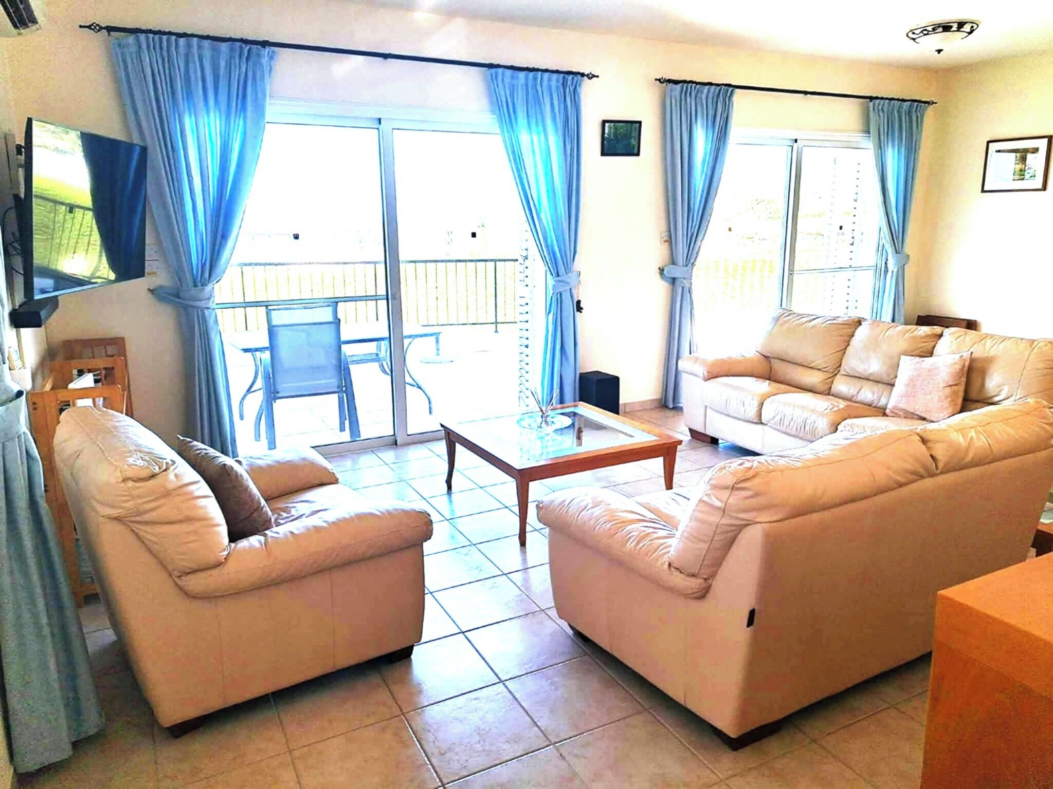 A living room with two sofas, a coffee table, and a TV. There are blue curtains and a sliding glass door leading to an outdoor area.