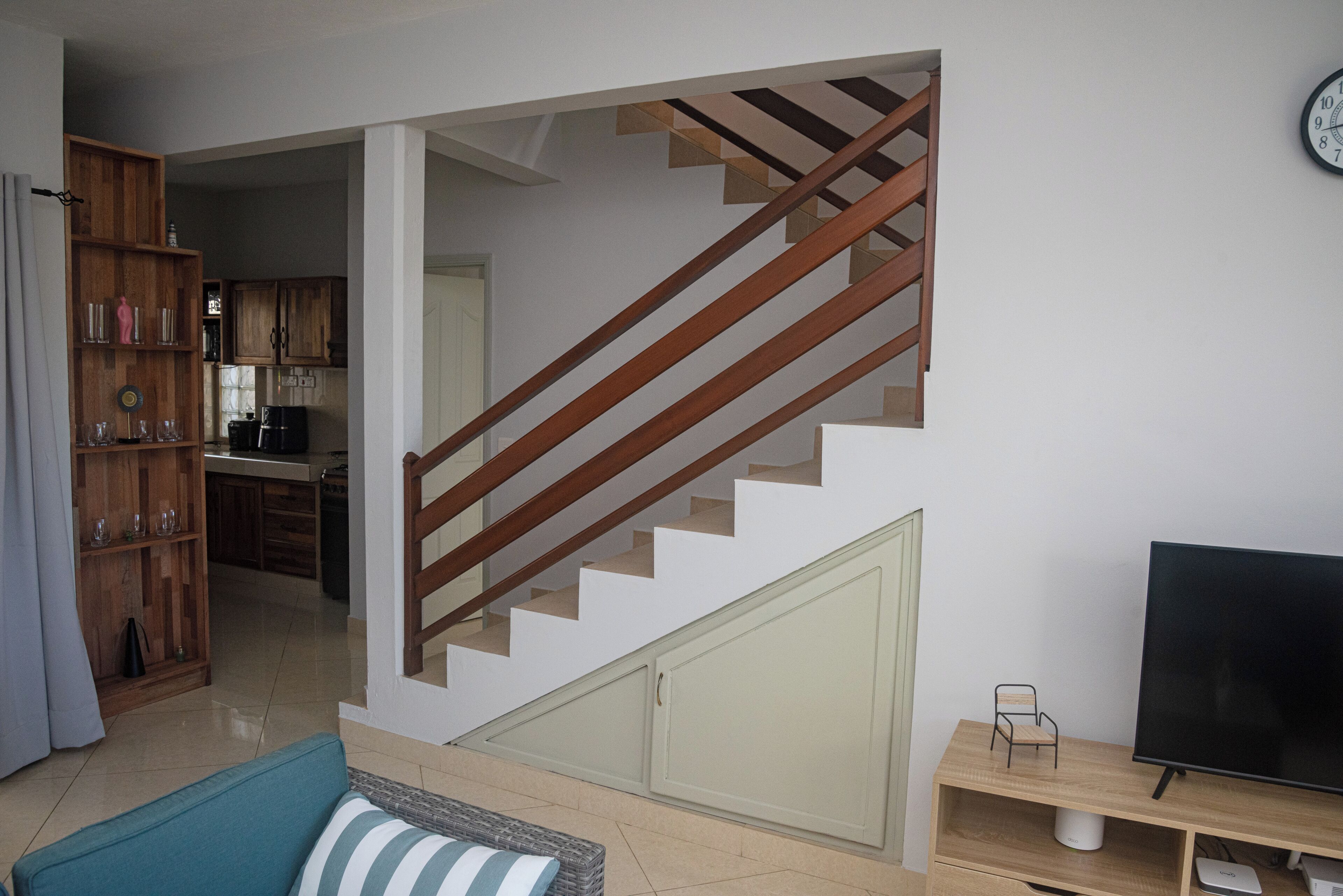 A staircase leading to a second floor, a dining area with wooden cabinets, a television on a wooden stand, and a sofa with a striped cushion.