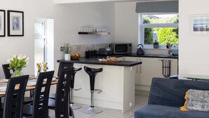 A modern kitchen with a central island, bar stools, a dining table with chairs, and a sofa. - West Wittering Beach House Garden Parking (Chichester)