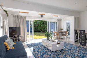 Una sala de estar moderna con sofá, mesa de centro y televisor, grandes puertas correderas de vidrio hacia un jardín, y un comedor con sillas y mesa.