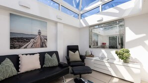 A modern living room with a black sofa, a black chair, a glass ceiling, a large painting, and a white console table with a plant. - West Wittering Beach House Garden Parking (Chichester)