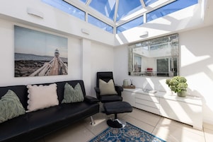 Una sala moderna con un sofá negro, una silla negra, techo de cristal, un cuadro grande y una consola blanca con una planta.