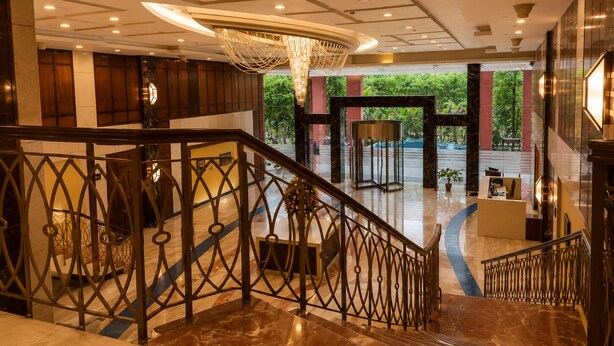 A hotel lobby with a marble staircase, a chandelier, and a view of greenery outside. - Bel La Monde Blu Chhattarpur (New Delhi)