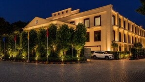A hotel with a paved courtyard, flagpoles, and a white car parked in front. - Bel La Monde Blu Chhattarpur (New Delhi)