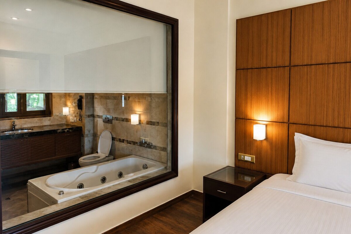 A hotel room with a large mirror, a wooden headboard, a bed, a nightstand, a bathroom with a bathtub, and a vanity with a sink and mirror.