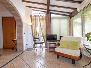 A living room with a sofa, a television, a lamp, a chair, and a window with blinds. - Rio del sole by Interhome (Montescudaio)
