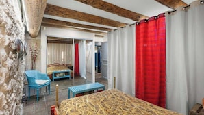 A room with exposed wooden beams, a bed with a straw-patterned cover, a blue chair, a bench, and red curtains.