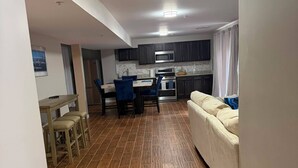 A modern kitchen with dark cabinets, a microwave, and a stove. There is a dining area with a table and chairs. The floor is covered with wooden tiles.