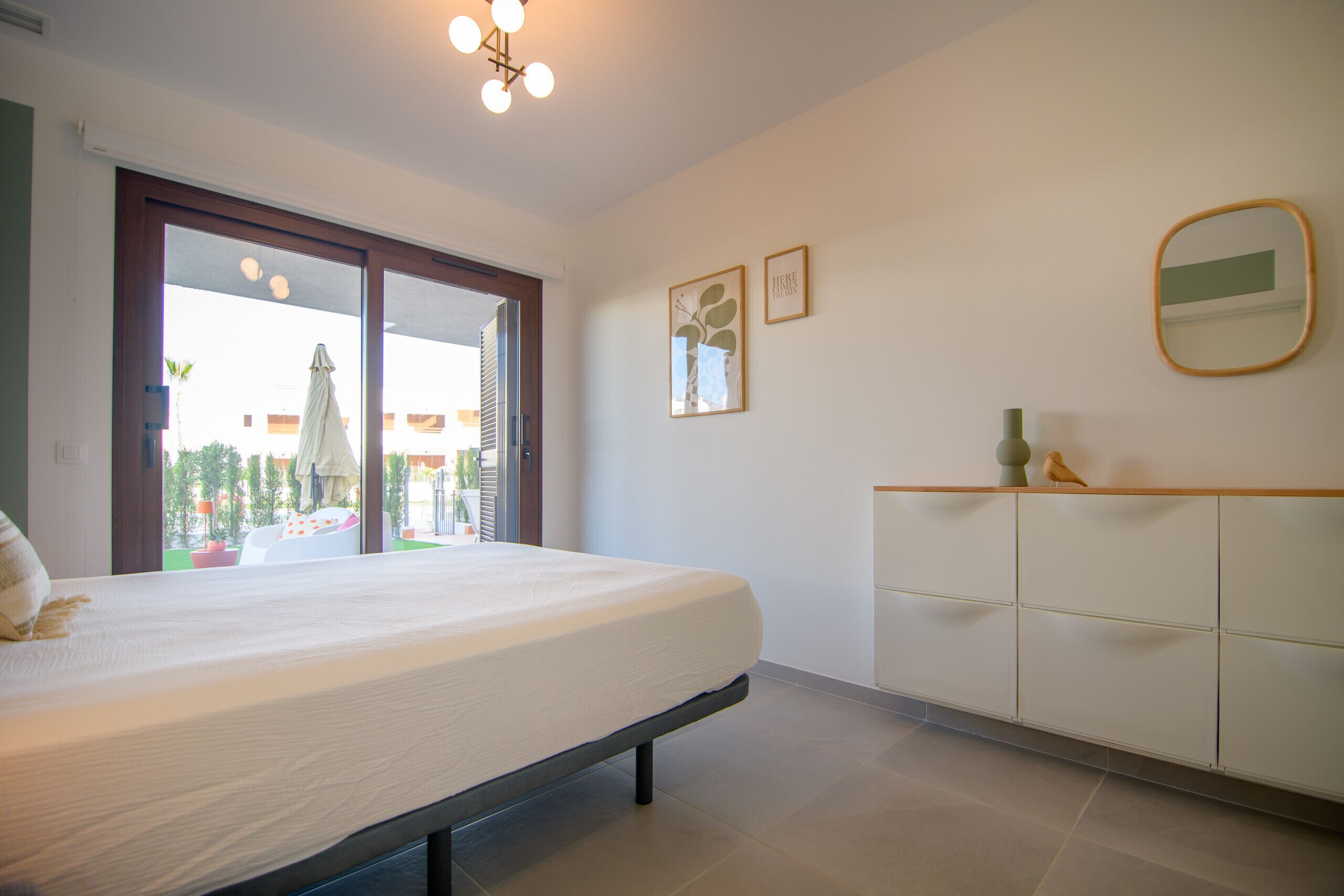 Quarto com cama, cômoda, espelho e vista através de uma porta de vidro.