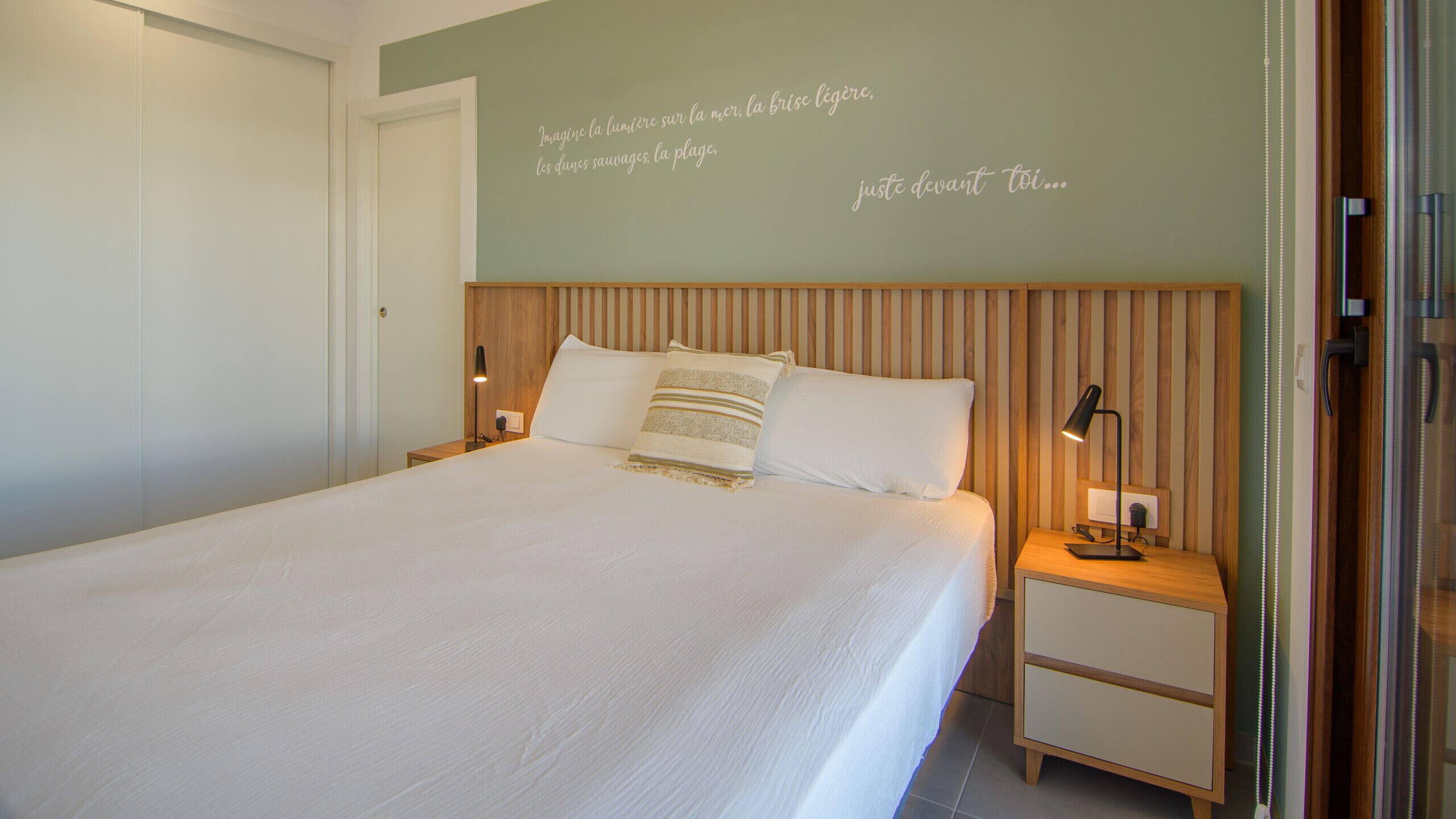 Un lit bien fait, agrémenté d’un coussin à rayures, une table de chevet avec une lampe et un mur orné d’une phrase décorative.