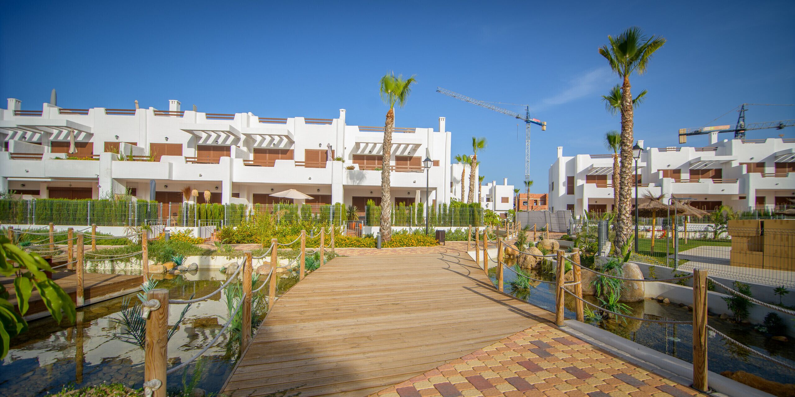 A resort with a wooden boardwalk leading to a pool area, surrounded by white buildings with balconies.