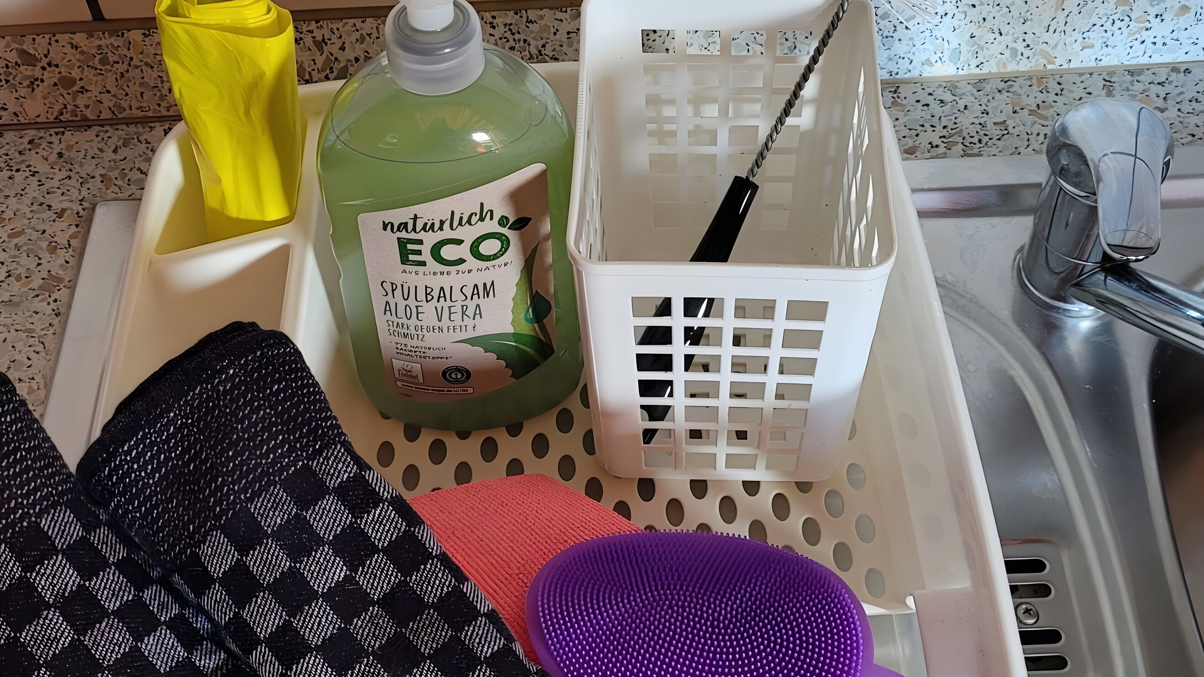 A kitchen area with a green bottle labeled 'natürlich ECO,' a white laundry basket, and a purple cleaning brush.