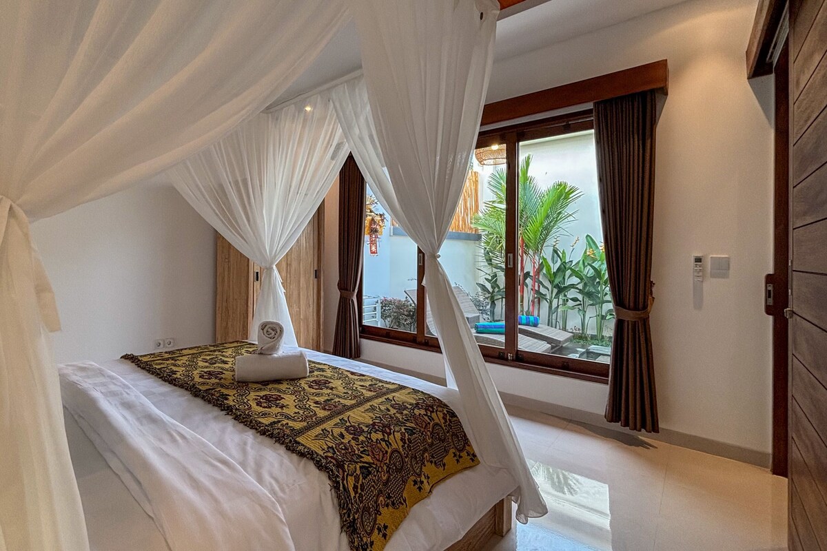 A bedroom with a canopy bed, a view of the outdoors, and a window with curtains.