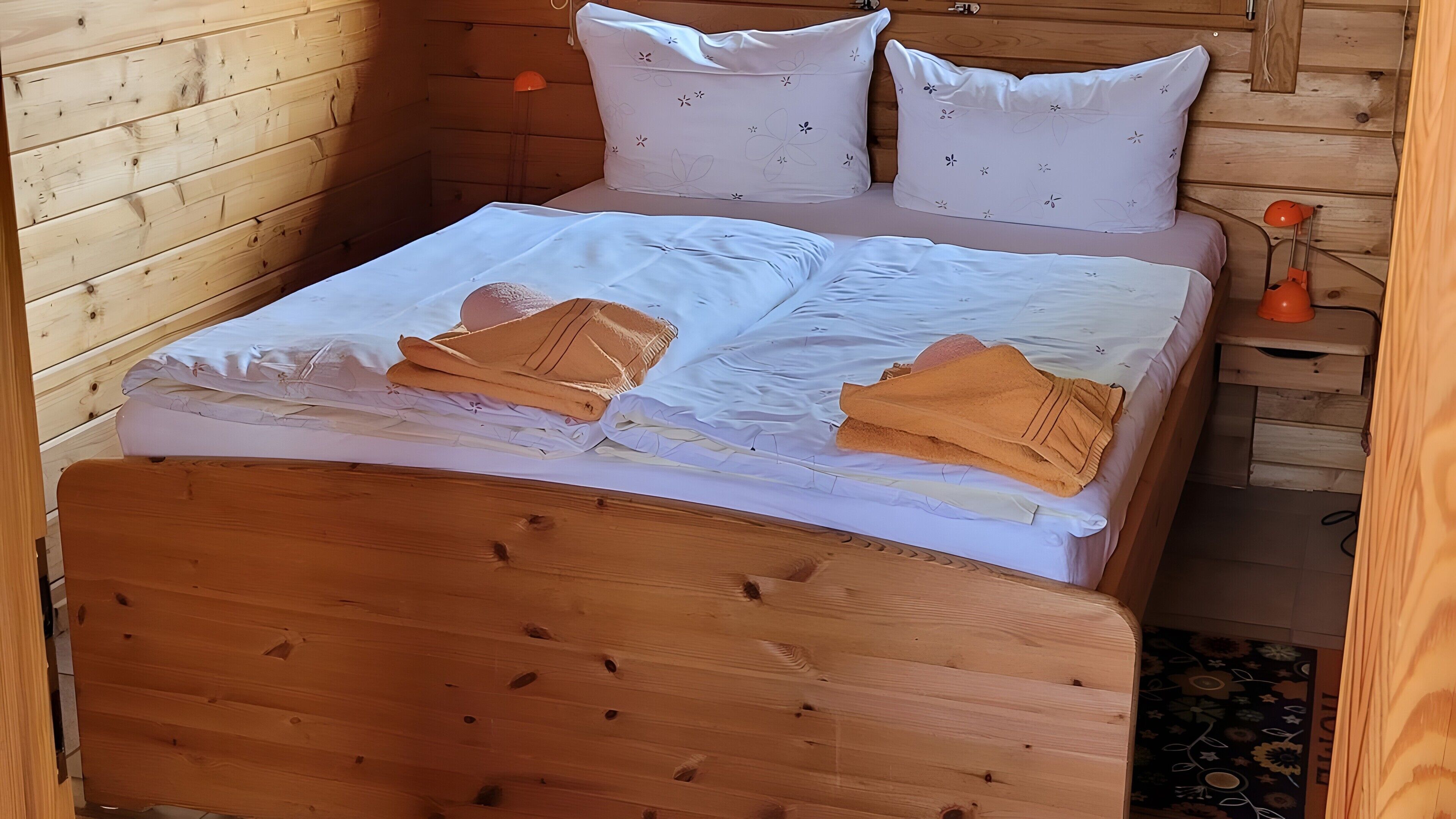 A wooden bed with white bedding and two pillows, a wooden headboard, and a small bedside table with an orange lamp.