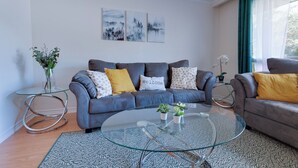 A modern living room with a grey sofa, a glass coffee table, and a small side table with a plant. - Stylish Home near Luxury Spa & GO Station (Whitby)