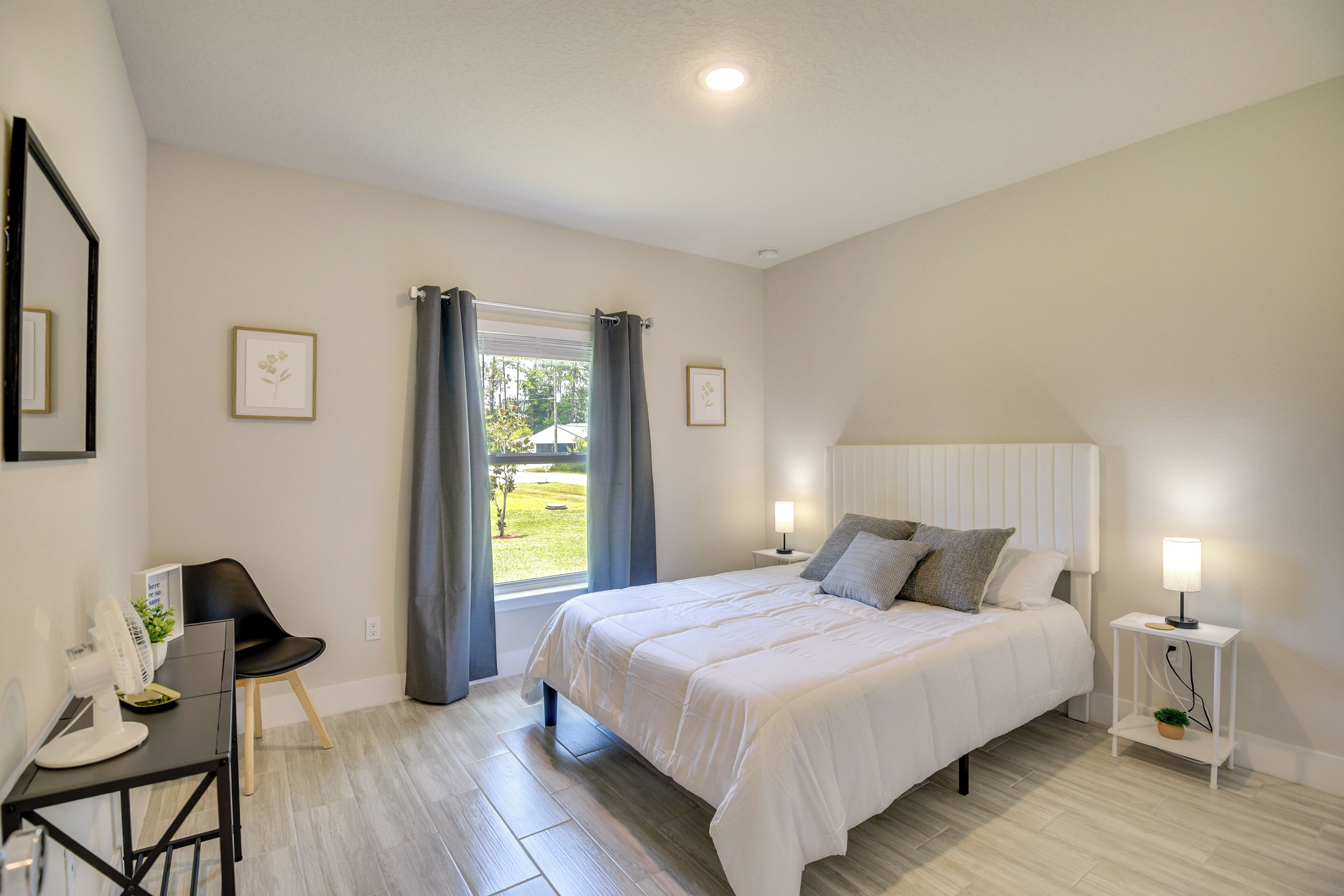 Quarto com cama, mesas de cabeceira, uma cadeira, escrivaninha e janela com cortinas.