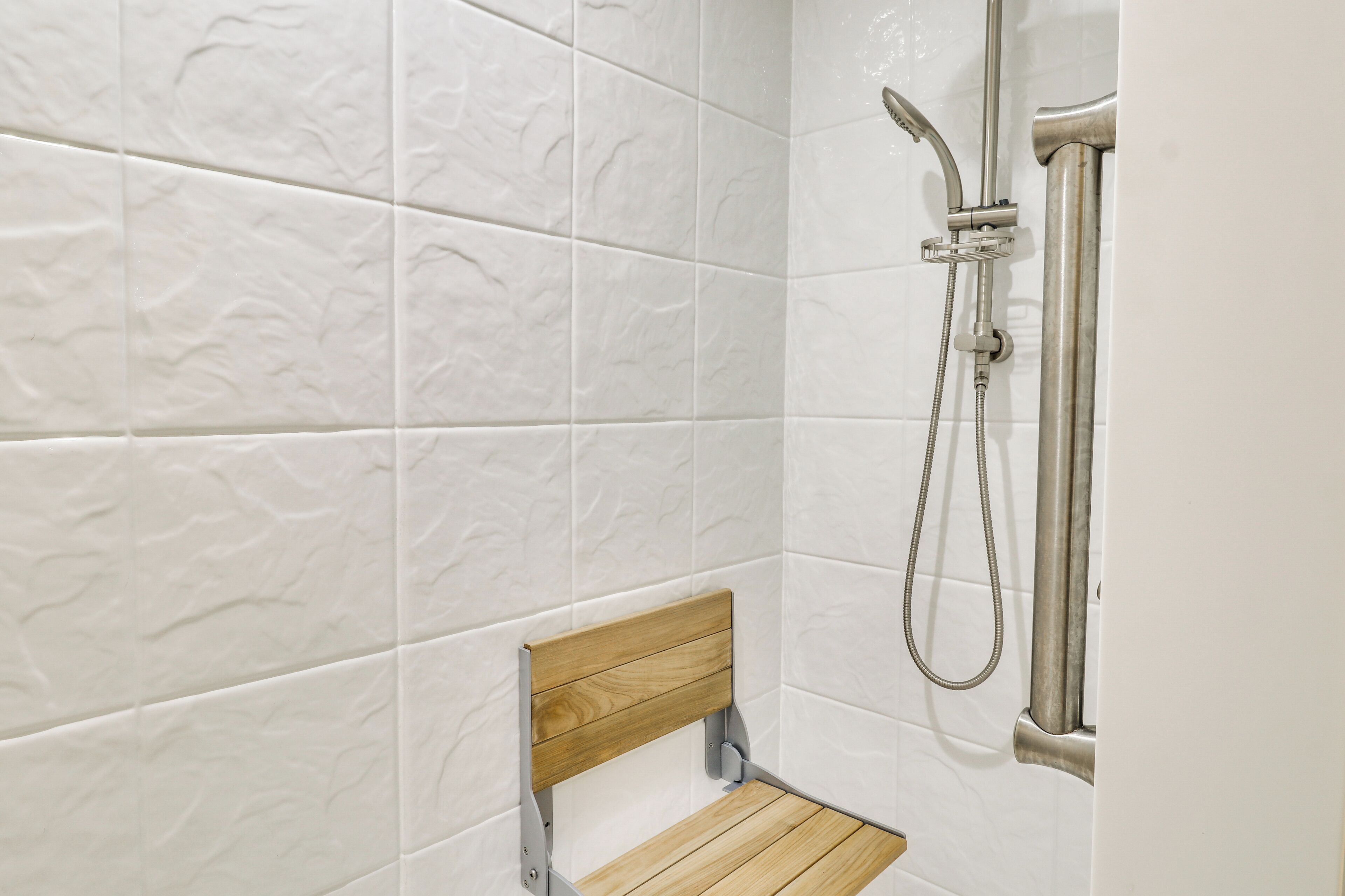A bathroom with white textured tiles, a wooden bench, and a shower with a handheld showerhead.