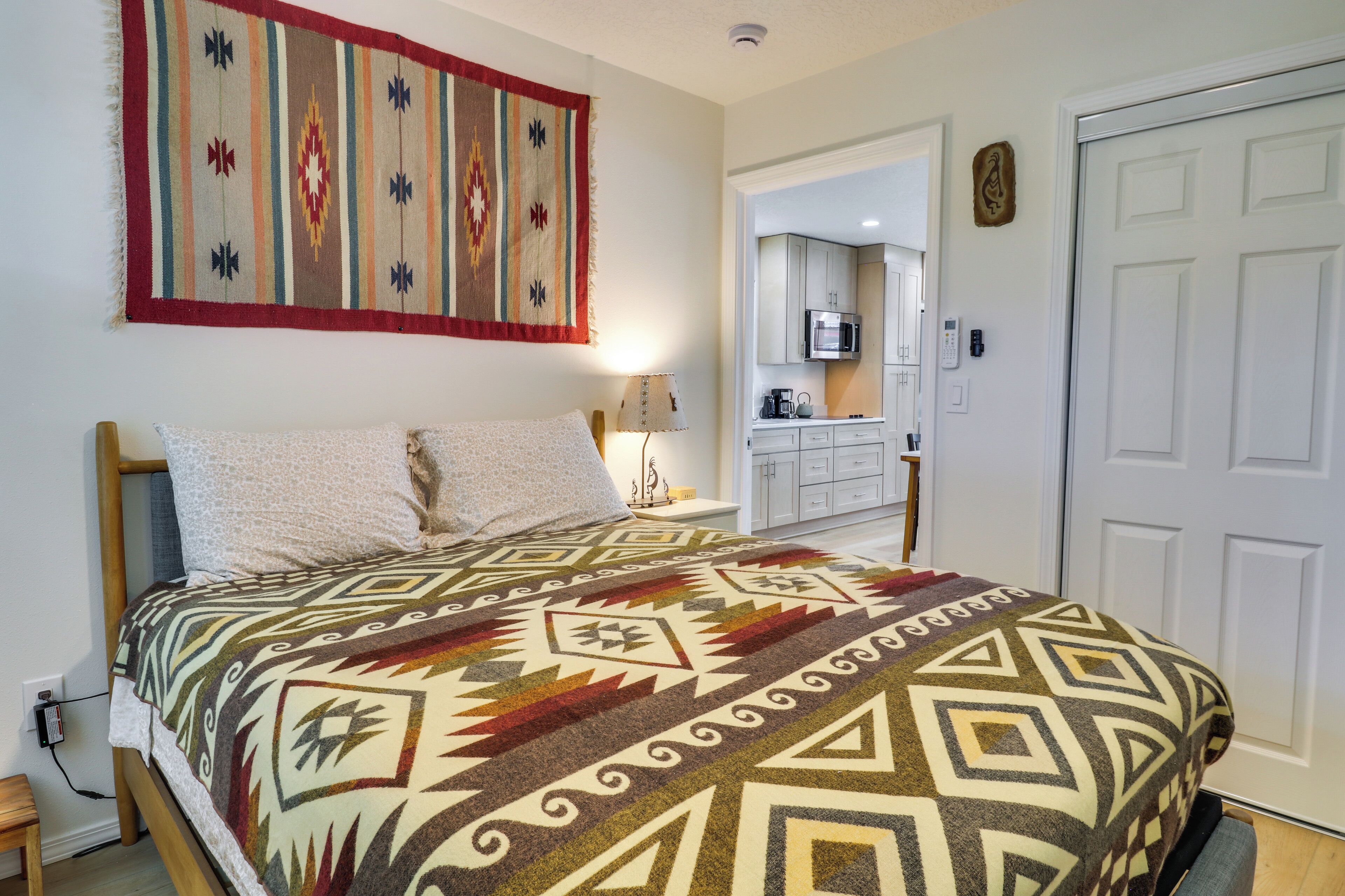 A bedroom with a bed, a nightstand, a lamp, a wall hanging, and a view into the kitchen.