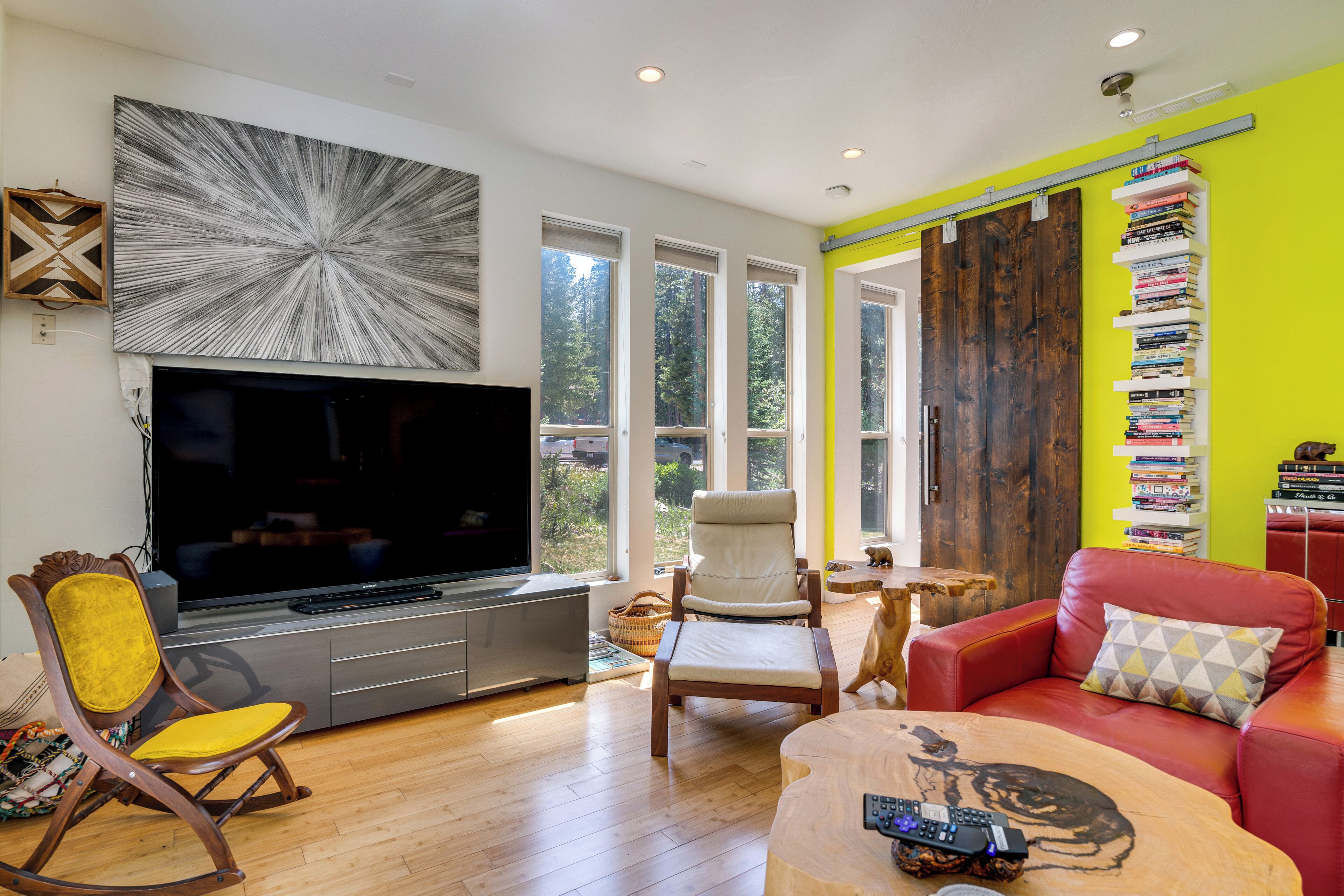 A living room with a flat-screen TV, wooden flooring, a red sofa, a yellow rocking chair, and a bookshelf.