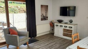 Una habitación con sofá, una mesa de comedor con sillas, televisor, armario y un ventanal con vistas al verdor.