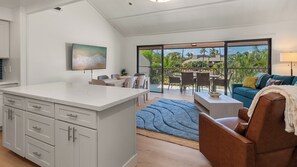 A modern kitchen with a marble countertop and white cabinetry, a dining area with a table and chairs, and a living area with a sofa and a view of the outdoors through large sliding doors. - Remodeled Spacious 3bd Across From Beach I Koa Resort 2H (Kihei)