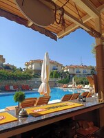 Zona junto a la piscina con sillones de descanso, un bar y vistas a edificios al fondo.