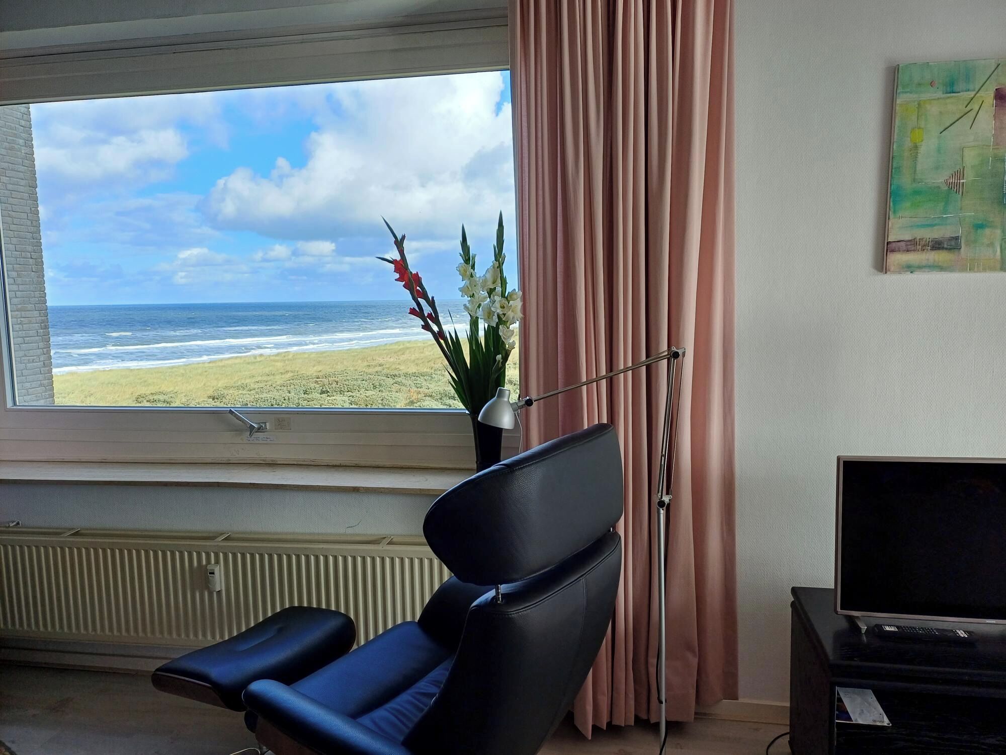 A room with a view of the beach, a black leather chair, a desk with a lamp, and a television.