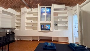 A room with a brick ceiling, white walls, a TV on a shelf, a massage table, and a door. - Elegant 5-day loft, Milan Dateo, tricolor (Milano)