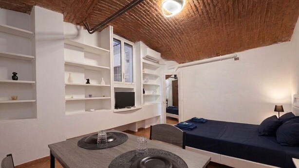 A compact room with a bed, a dining table with a chair, and built-in shelves with various items. - Elegant 5-day loft, Milan Dateo, tricolor (Milano)