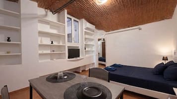 A compact room with a bed, a dining table with a chair, and built-in shelves with various items.