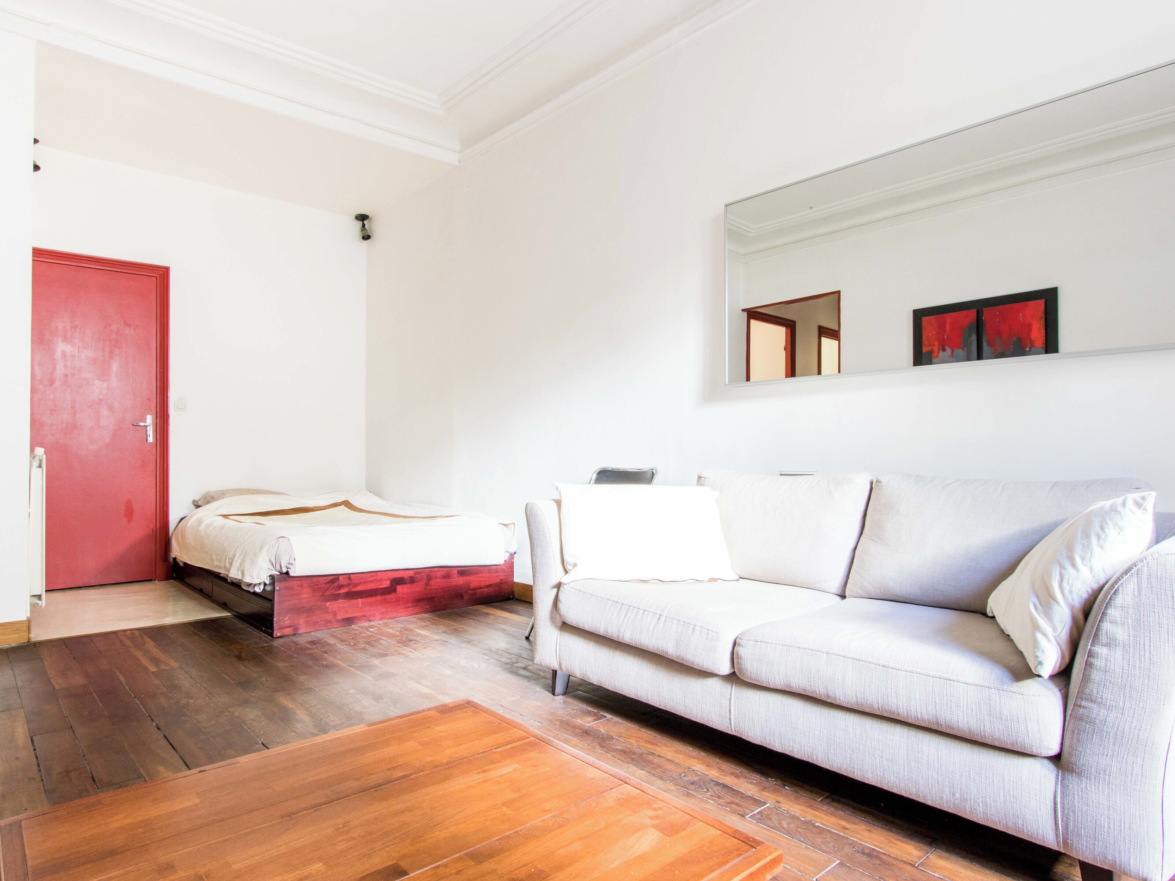 A room with a bed, a sofa, a red door, and a mirror on the wall.