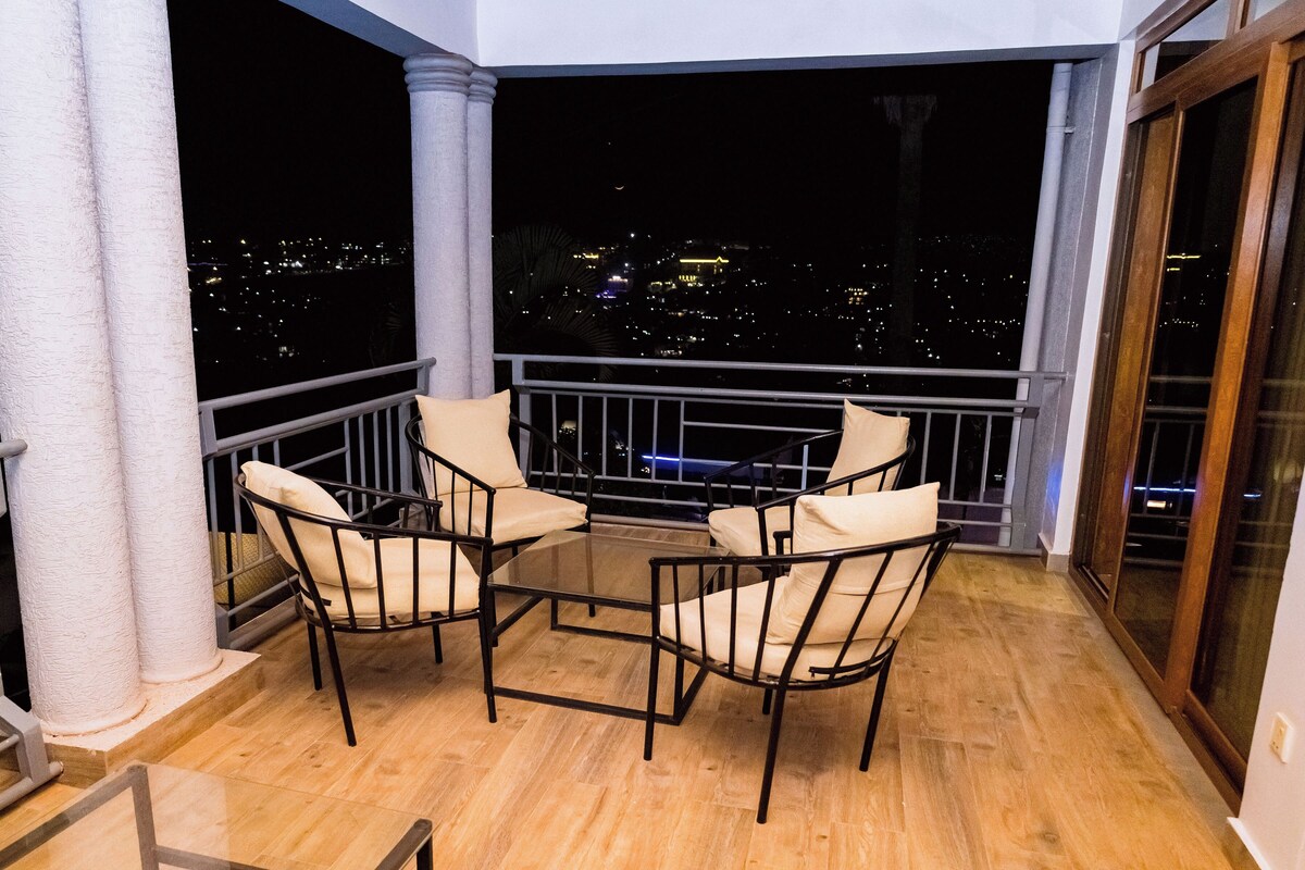 En balkon med et glastbord og fire stole, der giver udsigt over bybilledet om natten.