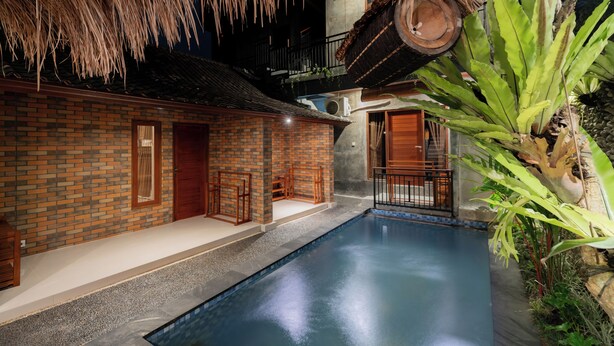 An indoor swimming pool with a brick wall, a thatched roof, and tropical plants. - Joni Ubud Hostel (Mas)