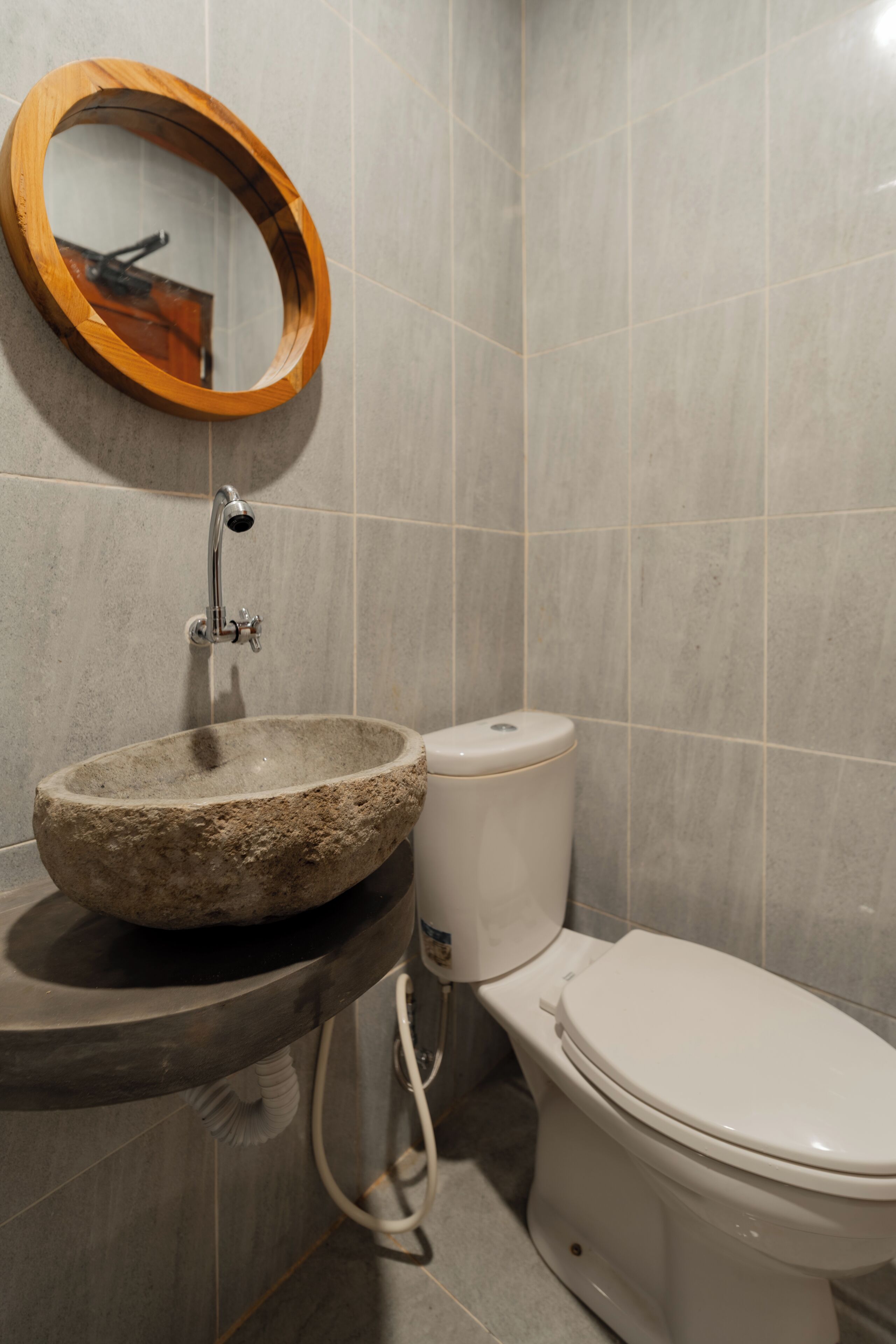 Ein Badezimmer mit einem Steintonne, einem spiegel mit Holzrahmen, einer weißen Toilette und grauen Fliesenwänden.