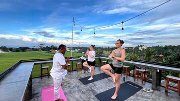 Un grupo de personas practicando yoga en colchonetas al aire libre, con vista a un paisaje verde y edificios al fondo.
