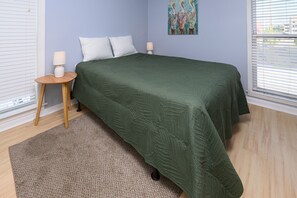 A bedroom with a bed, pillows, a bedside table with a lamp, a rug, and a painting on the wall. - 1st Floor, 2BR Oceanfront Condo, Shared Pool - Sand Dollar 103 (Murrells Inlet)