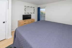 A bedroom with a bed, a television on a wooden dresser, a window with blinds, and a door. - 1st Floor, 2BR Oceanfront Condo, Shared Pool - Sand Dollar 103 (Murrells Inlet)