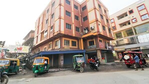 Un immeuble à plusieurs étages avec une entrée d’angle, une enseigne indiquant un épicerie, et plusieurs auto-rickshaws garés devant.