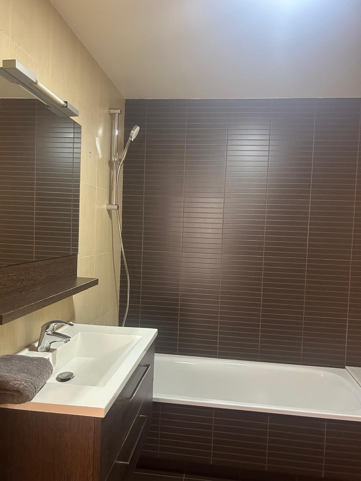 Un baño moderno con un lavabo blanco, un espejo, ducha y paredes con azulejos oscuros.