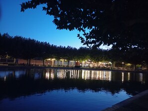 Escena tranquila de noche con un cuerpo de agua que refleja el entorno, árboles y un edificio iluminado al fondo.
