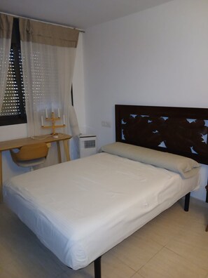 Habitación pequeña con cama individual, cabecera de madera, mesita de noche, una silla y ventana con cortinas.