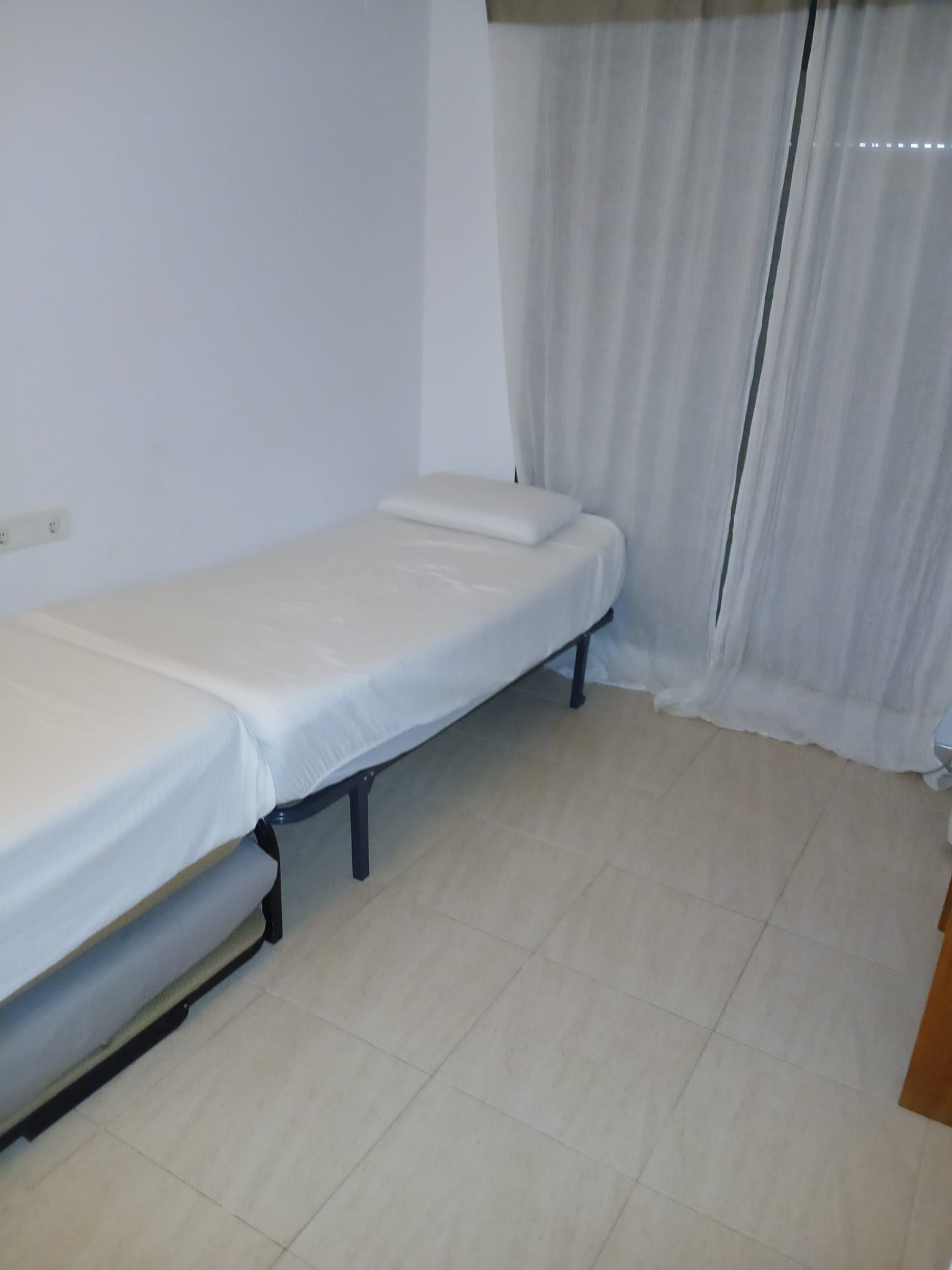Habitación con una cama individual, colchón y estructura de metal blancos, y una pared blanca sin adornos.