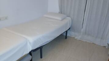 Habitación con una cama individual, colchón y estructura de metal blancos, y una pared blanca sin adornos.