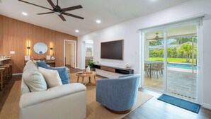 A modern living room with a ceiling fan, a flat-screen TV, a sofa, a coffee table, and a sliding glass door leading to an outdoor area with a pool. - Seminole Family Escape: Pool, Games & BBQ (Seminole)