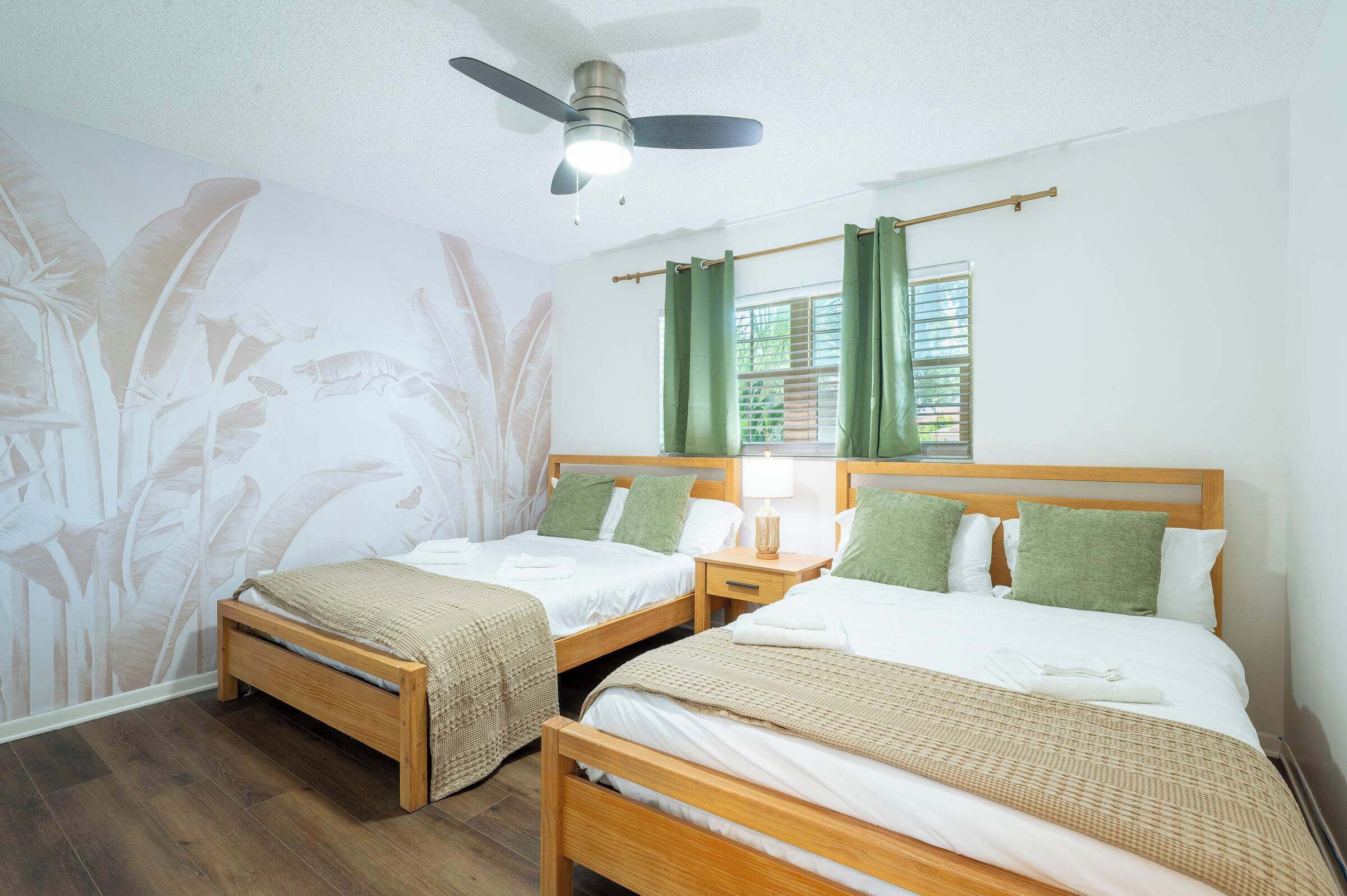 A hotel room with two wooden beds, a ceiling fan, a window with green curtains, and a wall mural.