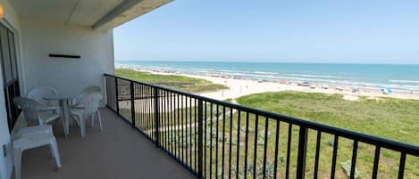 A balcony with a view of a beach, white plastic chairs, and a black railing.