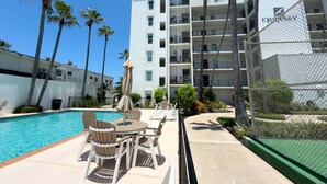 Un área de piscina con sillones de descanso y sombrillas, una cancha de tenis cercada y un edificio de varios pisos con balcones.
