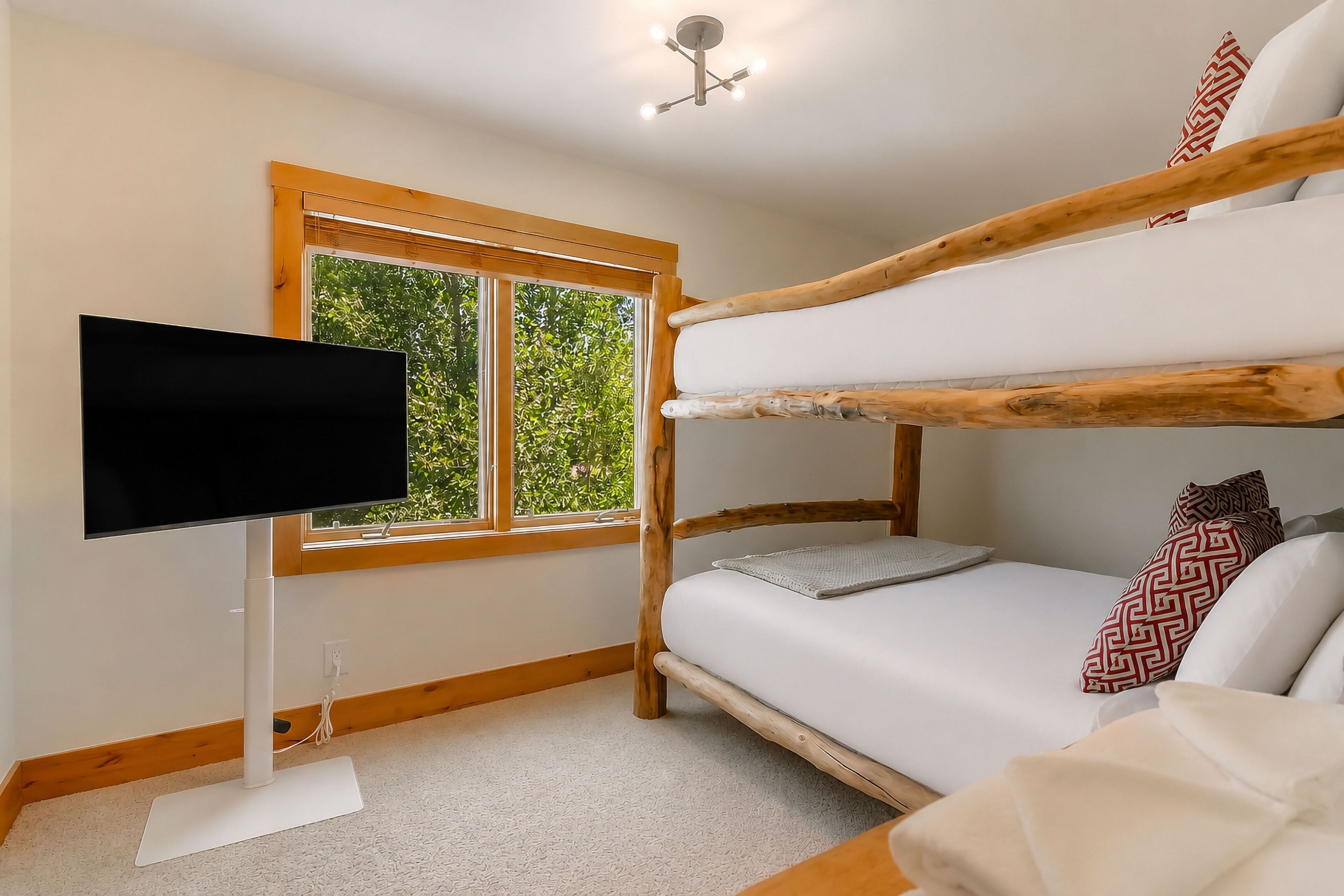 A bunk bed with white bedding, a television on a stand, and a window with a view of greenery.