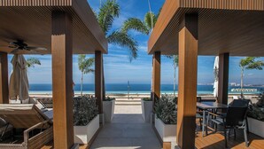 Ein überdachter Außenbereich mit Holzboden, Sitzgelegenheiten und Pflanzenkübeln mit Blick auf einen Strand und das Meer.