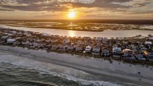 An aerial view of a coastal residential area with houses along the beachfront, a marina, and a sunset over the water. - Buoy Haus: Brand-New 2027 Inlet Front Escape w/Pool, Spa & Dock (Murrells Inlet)